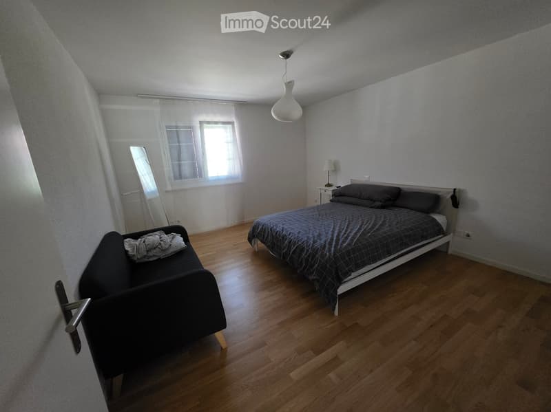 A modern bedroom with wooden flooring, a double bed with dark bedding, a small sofa, a mirror, and a nightstand with a lamp.