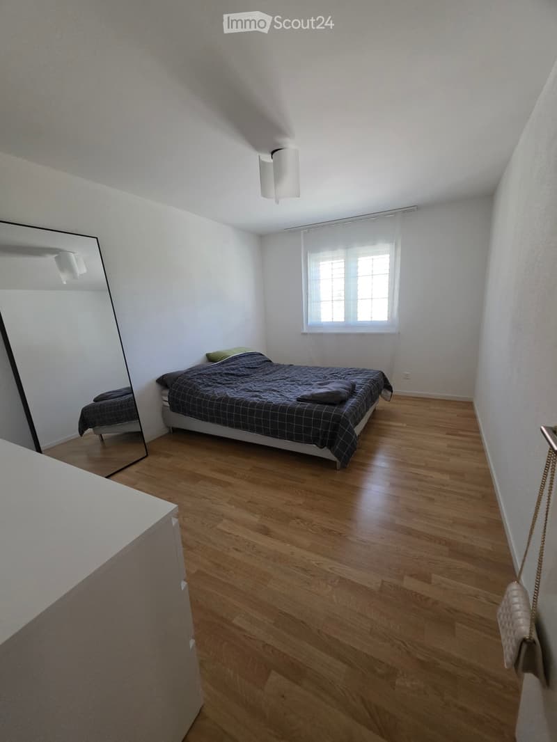 Bedroom with wooden flooring, a single bed with dark bedding, a large mirror, and a window with white curtains.