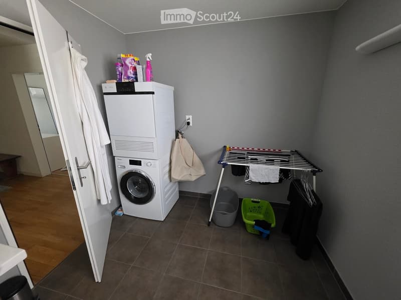 Small bathroom with a stacked washing machine and dryer, drying rack, handbag, and cleaning supplies.