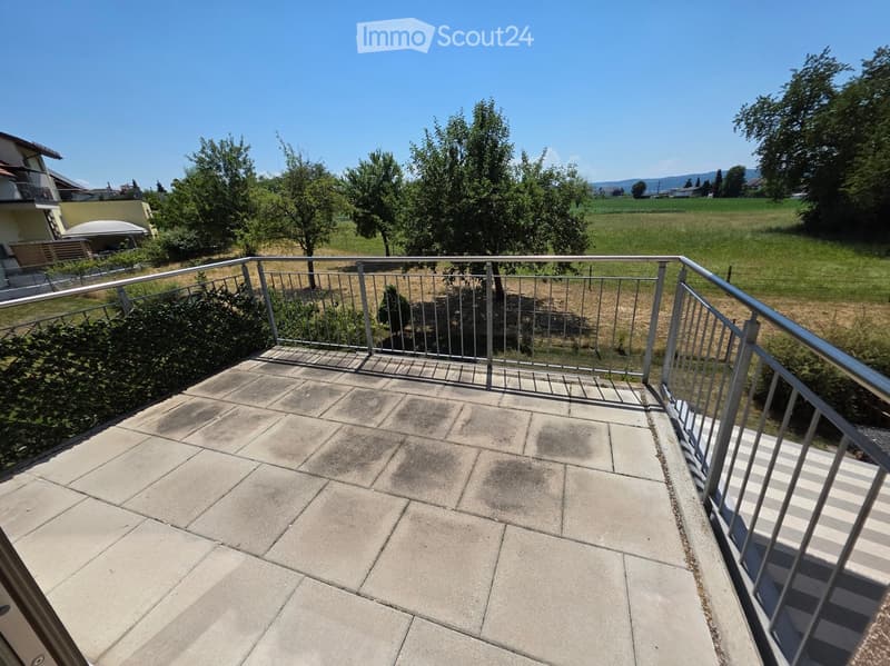 Balcony with concrete tiles, metal railing, and a view of a green field and trees.