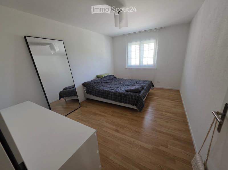 A modern bedroom with a wooden floor, a large mirror, a bed with a dark grey duvet and pillows, and a window with blinds.