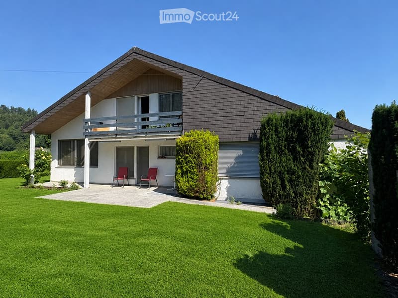 Two-story house with a modern design, featuring a slanted roof, large windows, and a balcony on the upper level. The front yard includes a well-maintained lawn, trimmed hedges, and a small patio area with two red chairs and a table.