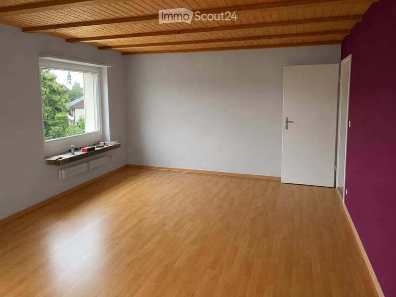 Empty room with wooden flooring, white walls, and a large window with a view of greenery. There is a white door on the right side and a small shelf with a few items on the left side.
