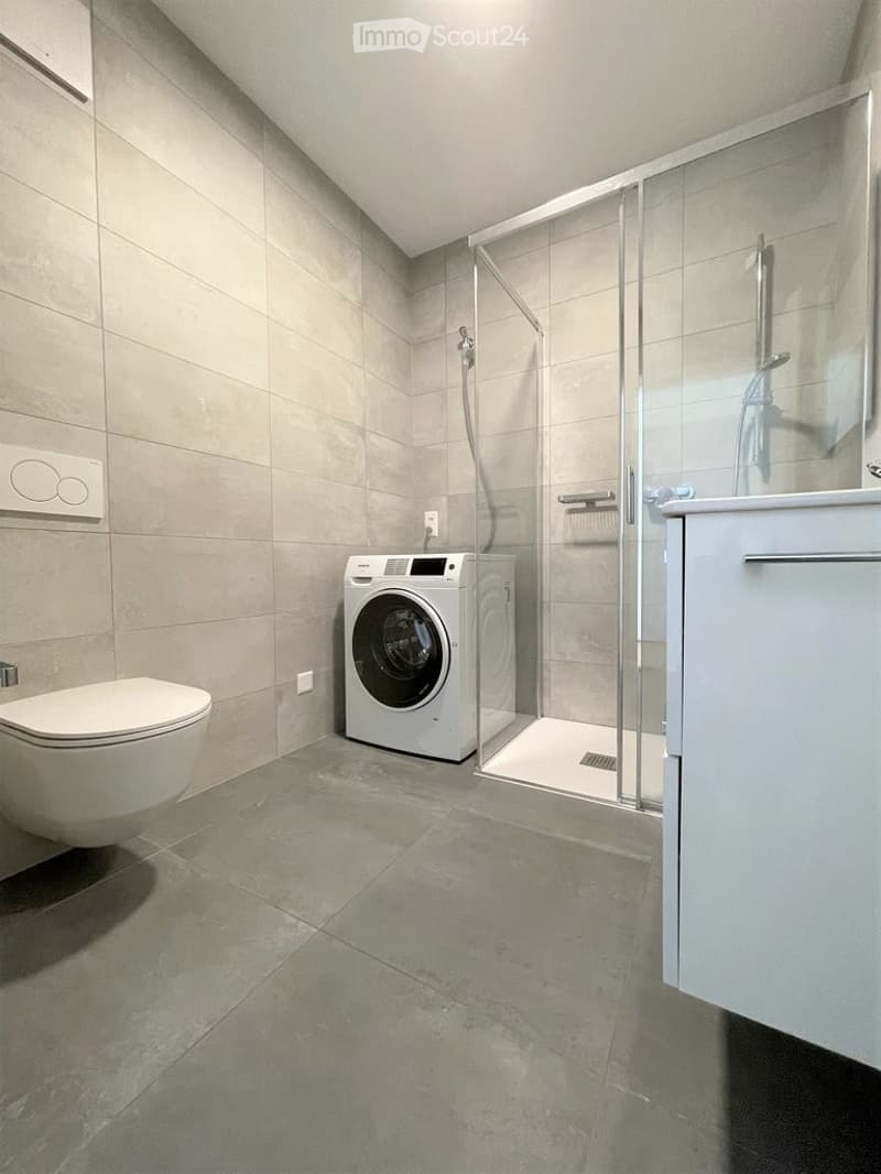 Modern bathroom with a toilet, washing machine, glass-enclosed shower stall, and white cabinet.