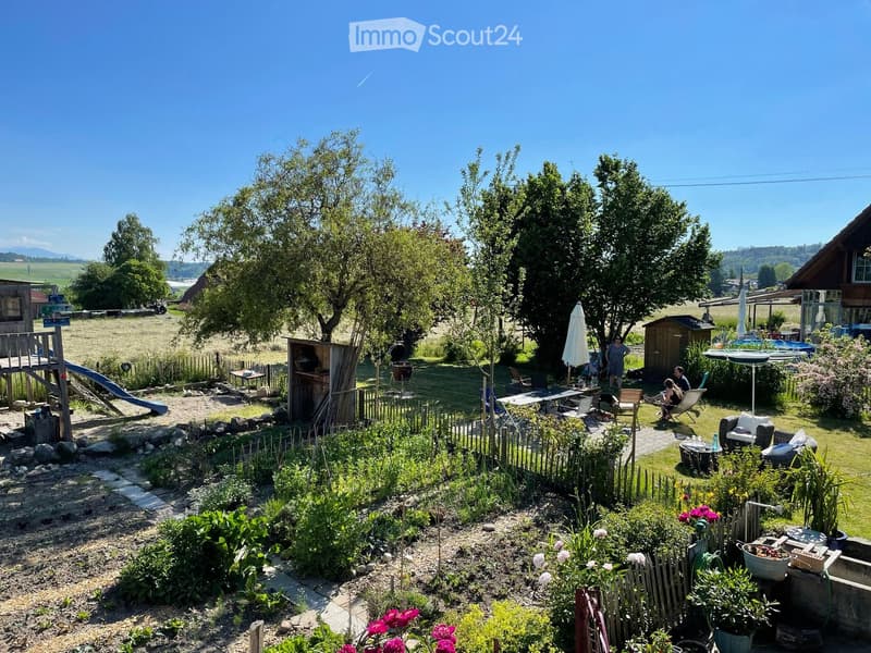 A spacious garden with a mix of seating areas, a small play structure, and a well-maintained flower bed. The area includes outdoor furniture, a grill, and a scenic view of the countryside.