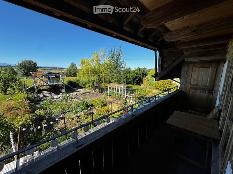 Wooden balcony with a table and chair, overlooking a garden and countryside landscape.