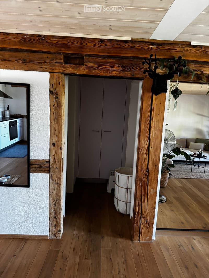 A wooden-floored hallway with exposed wooden beams and a large open doorway leading to a living area with a sofa, coffee table, and plants. A kitchen area is visible to the left with cabinets and appliances.