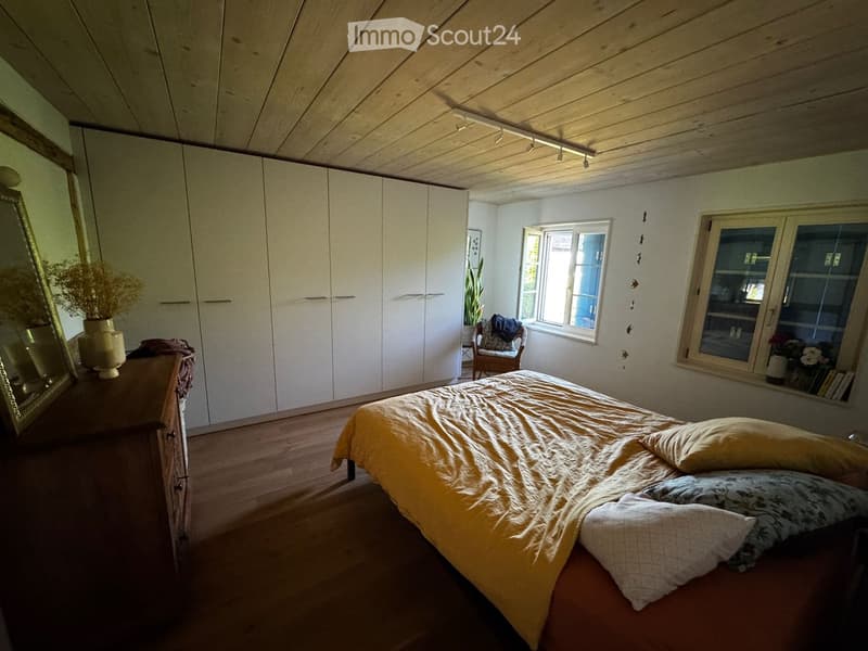 Bright bedroom with wooden floors, large windows, a neatly made bed with yellow bedding, a wardrobe, a mirror, a chair, and a vase with dried flowers on a dresser.