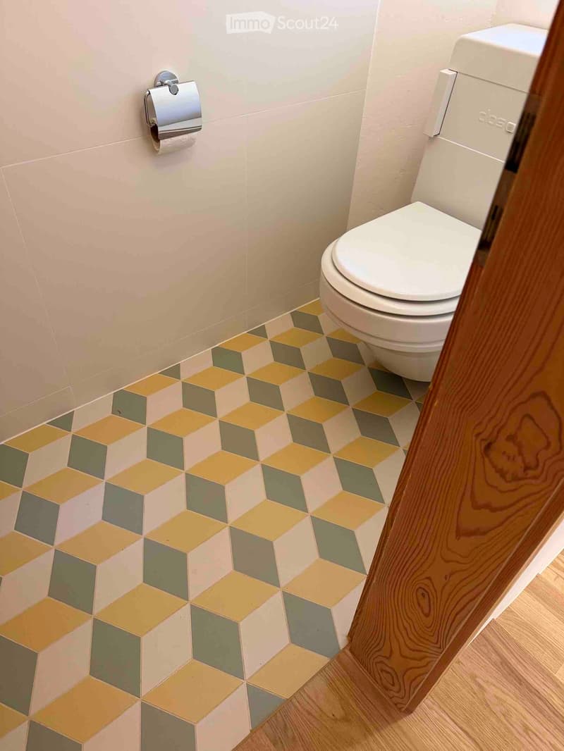 Bathroom with a white toilet, beige walls, and a colorful hexagonal tile floor in shades of yellow, green, and gray.