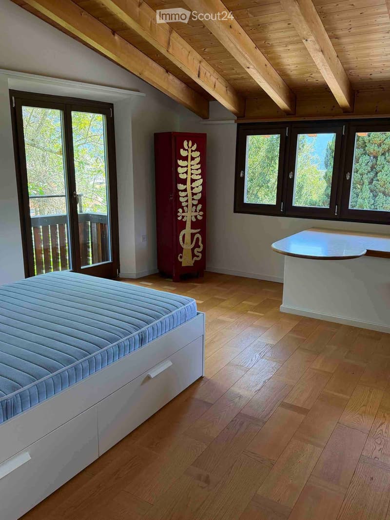 Bright bedroom with wooden floors, a large bed, a red wardrobe with a leaf design, and a round table near the window.