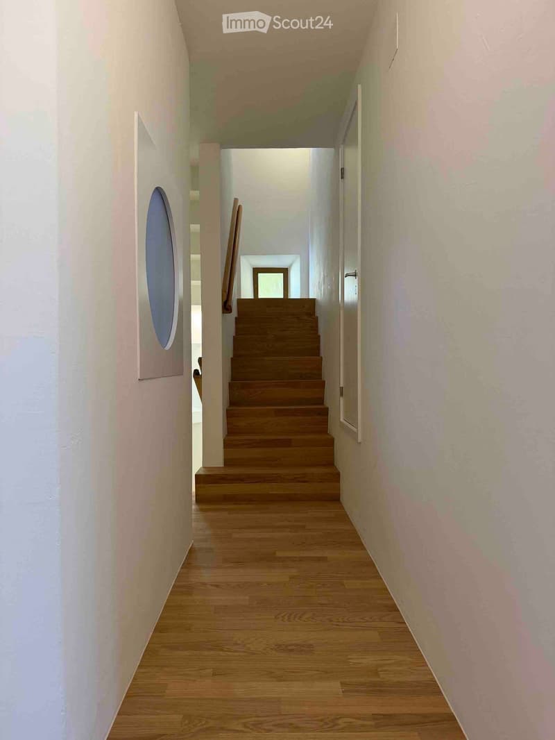 A modern hallway with wooden flooring, white walls, and a wooden staircase leading upwards. The hallway features a circular artwork on the left wall and a small open doorway at the end.