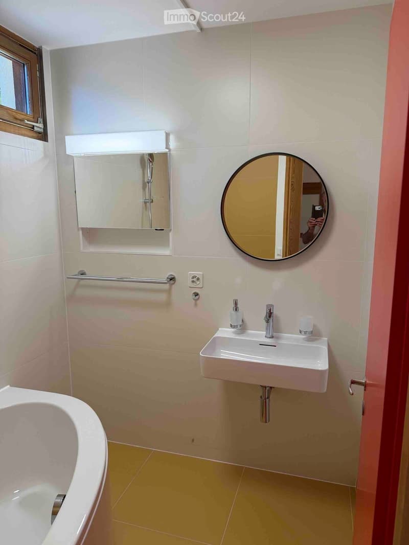 Modern bathroom with a white bathtub, a white sink with a modern faucet, a round mirror, a built-in medicine cabinet, and a towel rack.