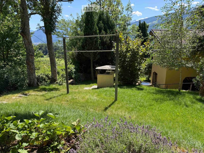A well-maintained garden with green grass, lavender plants, trees, and a small yellow shed in the background, with mountains visible in the distance.