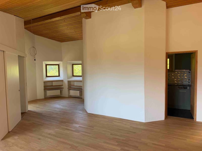 Empty room with wooden flooring, white walls, and exposed wooden ceiling beams. Two small windows with wooden frames and built-in shelving units are visible, along with a doorway leading to a kitchen area.