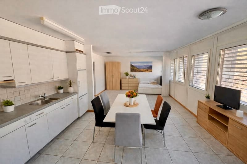 Bright, modern open-plan living and dining area with a fully equipped kitchen, dining table with chairs, TV stand, and a separate bedroom area with a bed and wardrobe.