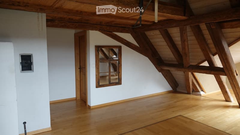 Empty attic room with wooden beams, exposed wooden ceiling, wooden floor, and a small window with wooden frame.