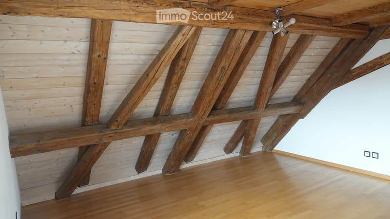Empty room with exposed wooden beams and a wooden floor, featuring a simple white wall and a small light fixture on the ceiling.