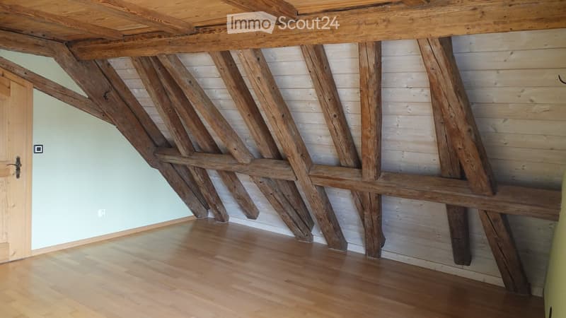 Empty room with exposed wooden beams and a wooden floor, featuring a simple wooden door and light green wall.