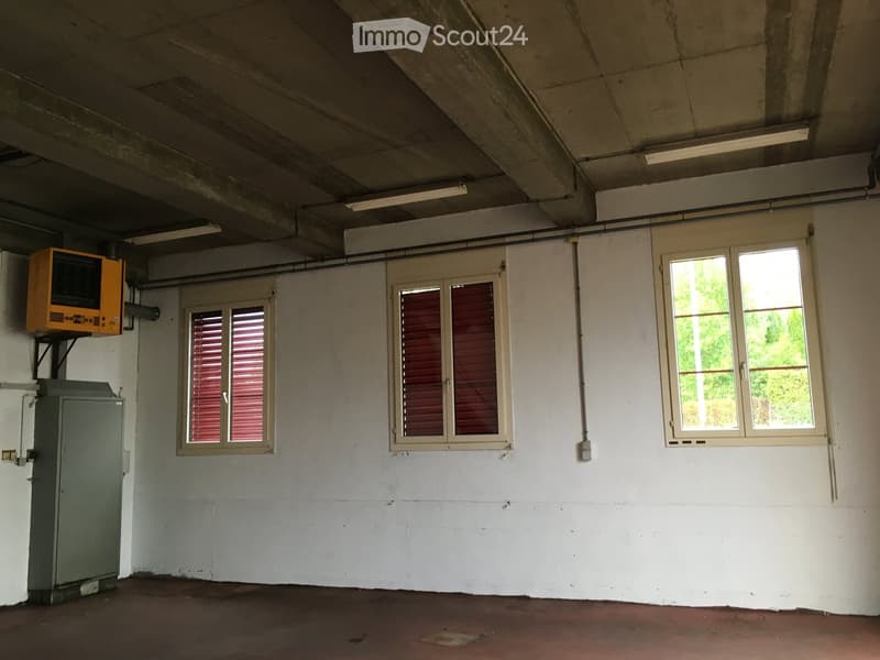 Empty room with white walls, concrete ceiling, and three windows with blinds. Features a grey electrical box and an orange air conditioning unit mounted on the wall.