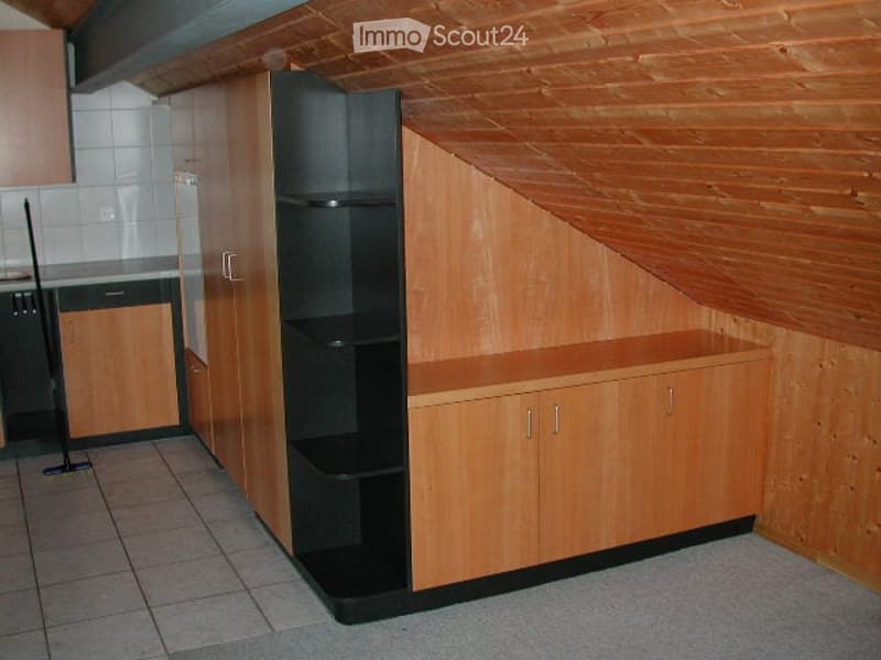 A small, well-lit room with wooden walls and a slanted ceiling, featuring built-in wooden cabinets and a black shelving unit.