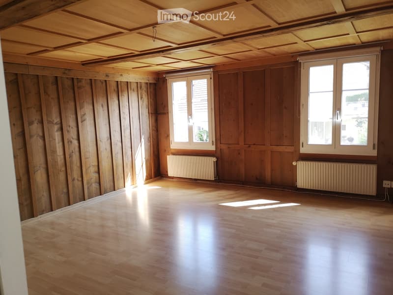 Empty room with wooden walls, wooden ceiling, wooden floor, and two windows with radiators beneath them.