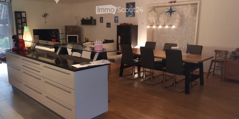 Modern kitchen and dining area with a central island, dining table, and chairs. The space features a sleek design with a glass table, wooden flooring, and contemporary decor.