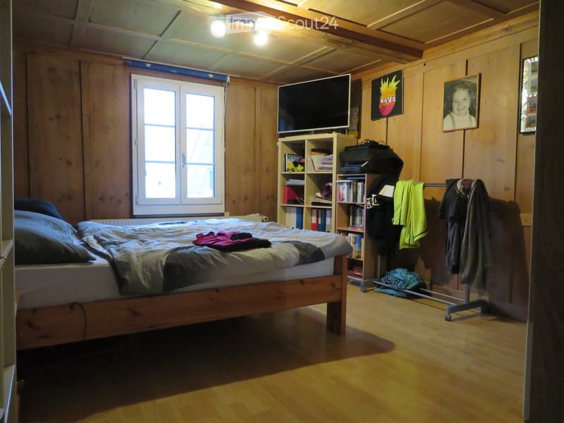Cozy bedroom with wooden walls and ceiling, featuring a double bed, a shelf with books and items, a television mounted on the wall, and a clothing rack with jackets and shirts.