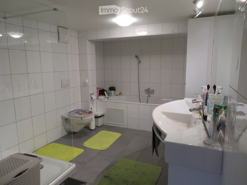 Modern bathroom with white tiles, a toilet, a sink with toothbrush holder, a bathtub with a shower hose, and green bath mats on the floor.