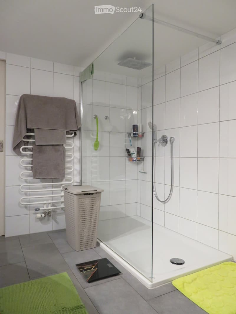 Modern bathroom with a glass-enclosed shower, tiled walls, towel rack with towels, shower shelf, bathroom scale, and a laundry basket.