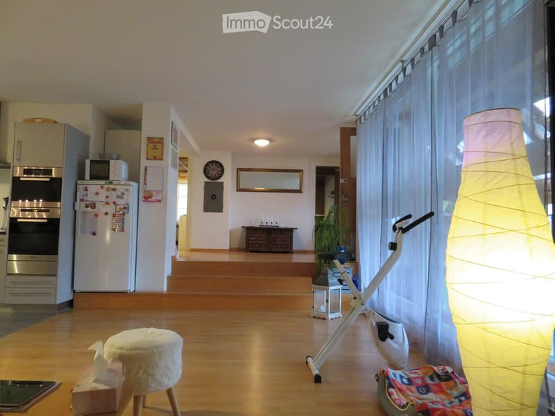 Bright, open living space with wooden flooring, large windows with white curtains, a tall floor lamp, a white exercise bike, a small stool, and a kitchen area with stainless steel appliances.