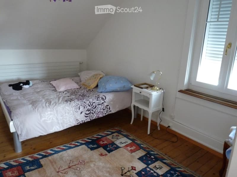 Cozy attic bedroom with wooden floors, a bed with colorful pillows, a nightstand with a lamp, and a patterned rug.