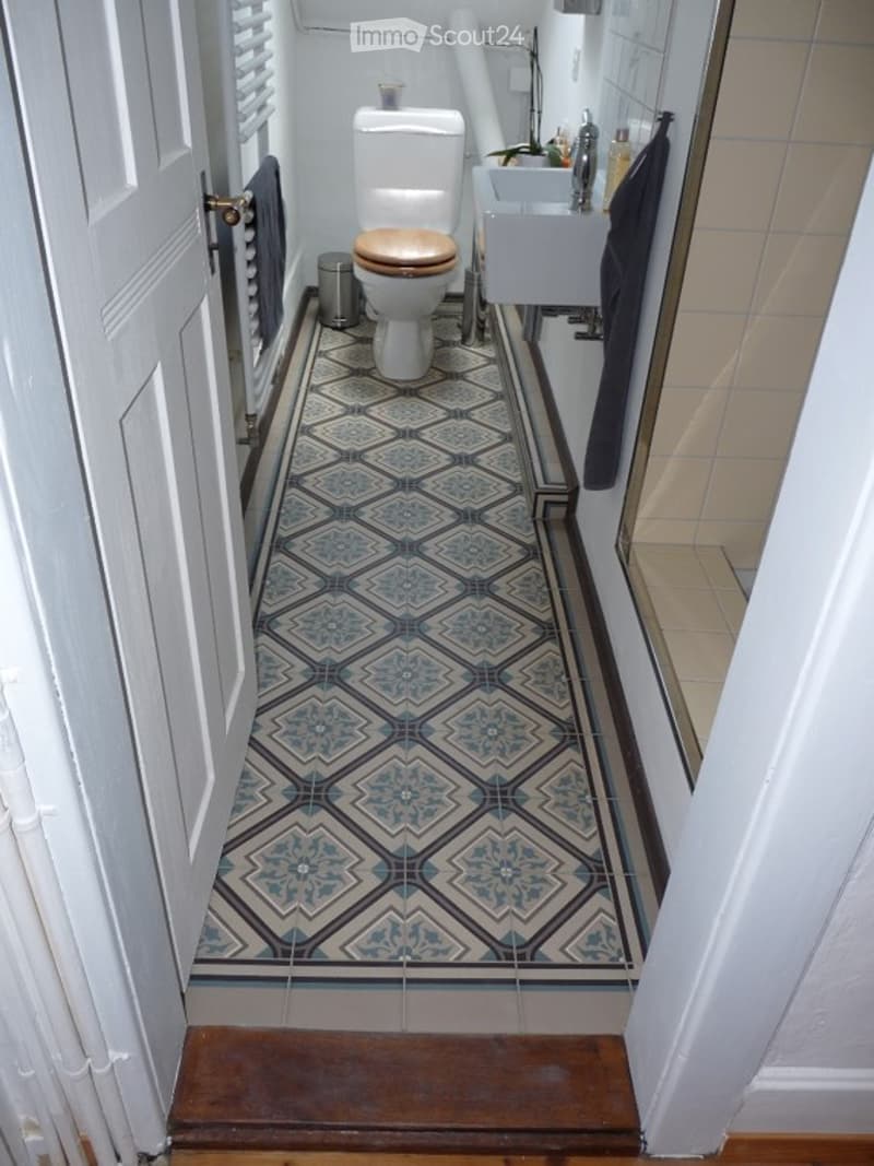 Modern bathroom with blue and white patterned tiles, a toilet with a wooden seat, a white sink, and a towel hanging on the wall.