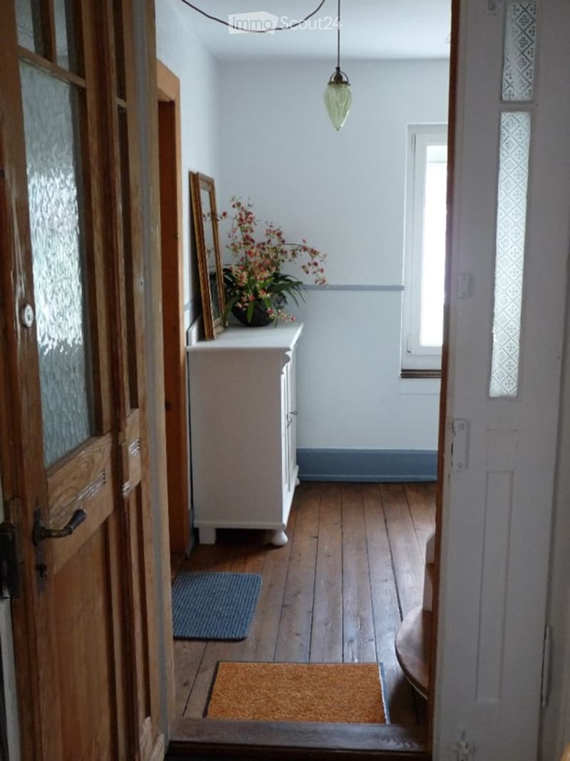 A narrow hallway with wooden flooring, white walls, and a small cabinet holding a flower arrangement and picture frame. Two doormats are placed on the floor near the entrance.