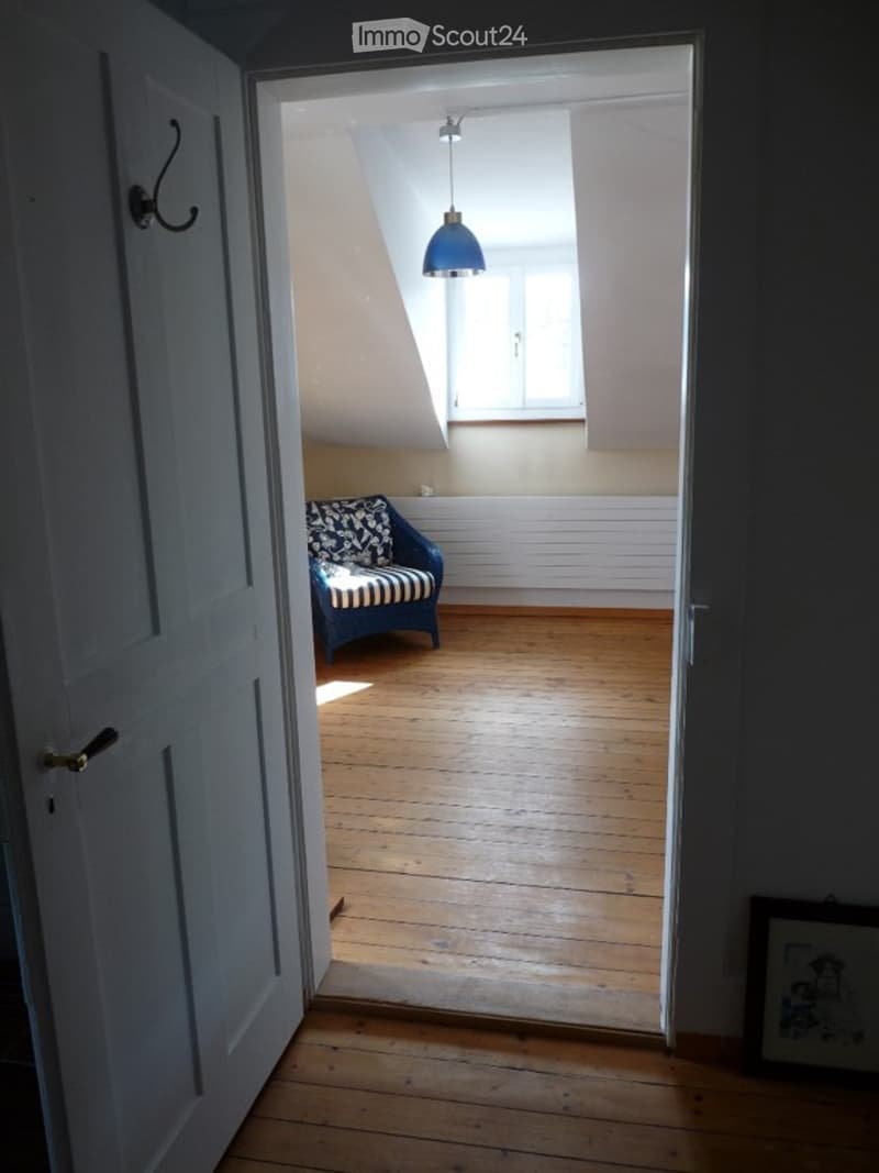 A small, bright room with wooden flooring and white walls. A blue and white striped sofa is positioned near a window, and a pendant light hangs from the ceiling.