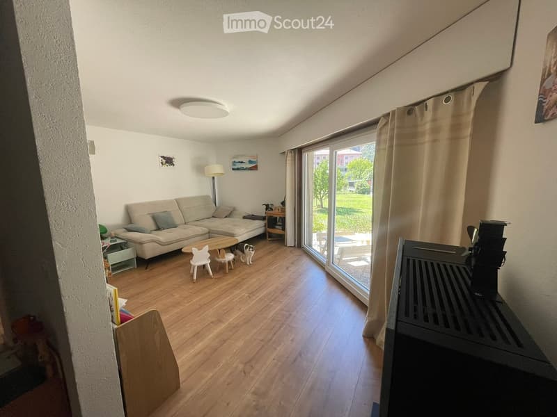 Bright living room with wooden flooring, a sectional sofa, coffee table, small chairs, and a cat. Large window with curtains offers a view of the garden.
