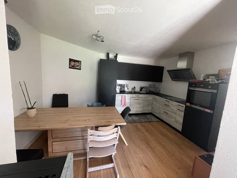 Modern kitchen with dark cabinets, light wooden table, white chairs, and various appliances including a refrigerator, oven, and toaster. The space features a potted plant and a clock on the wall.
