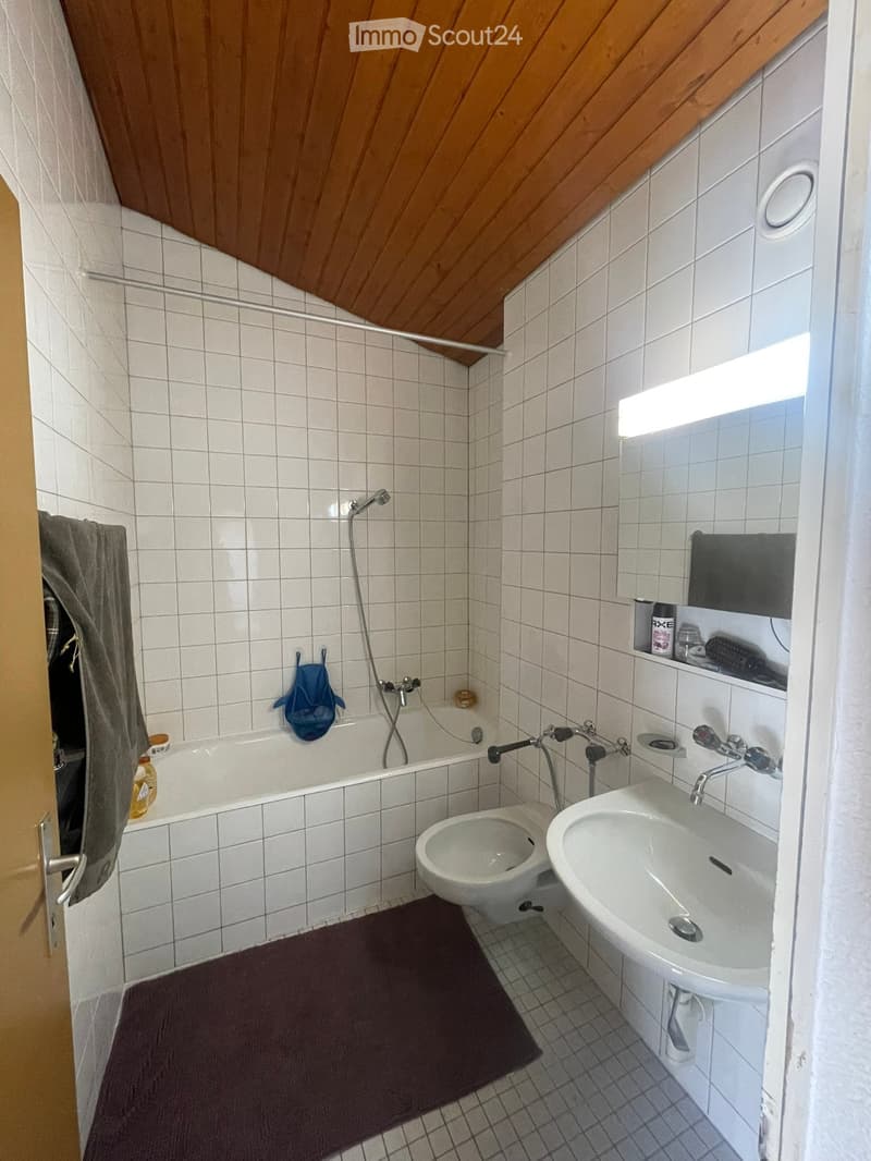 Bathroom with white tiled walls and floor, featuring a bathtub, toilet, sink, mirror, and a towel hanging on the door.