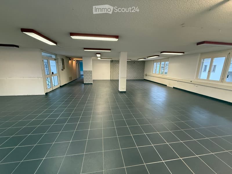 Empty room with gray tiles, white walls, fluorescent lighting, doors at both ends, and windows.