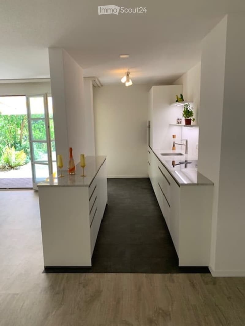Modern, minimalist kitchen with white cabinets, a central island, and a sleek countertop. Features a built-in sink with a modern faucet, and decorative wine glasses on the island.