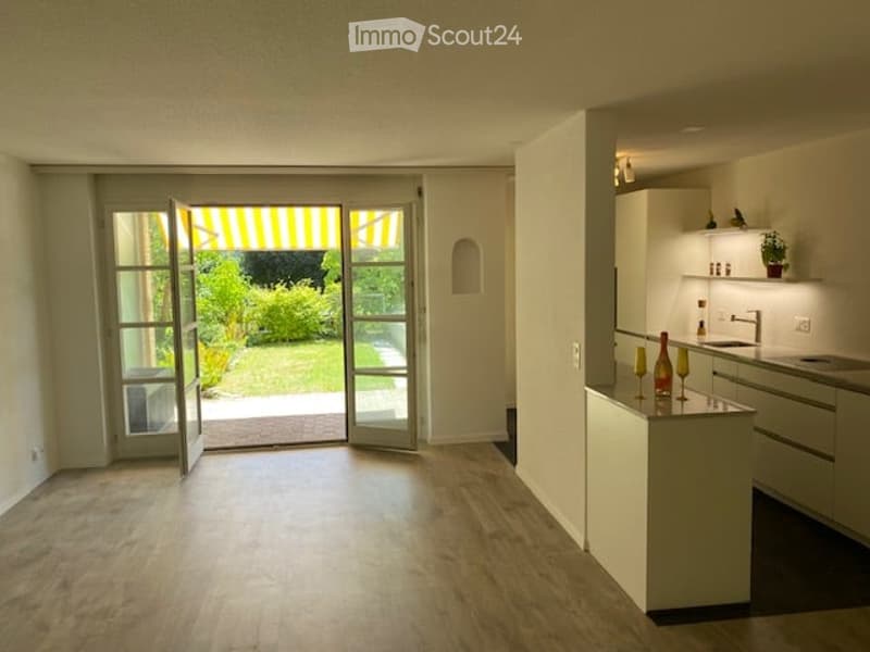 Bright, modern kitchen with white cabinets, a central island, and a large open doorway leading to a garden. Wine bottles and glasses are placed on the island, and a faucet is visible on the counter.