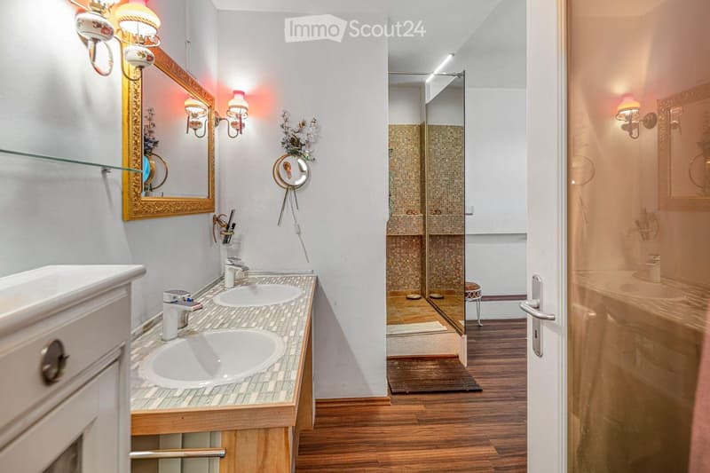 Bathroom with double sink, mirror, shower, wooden floor, wall-mounted lamps, and a glass door.