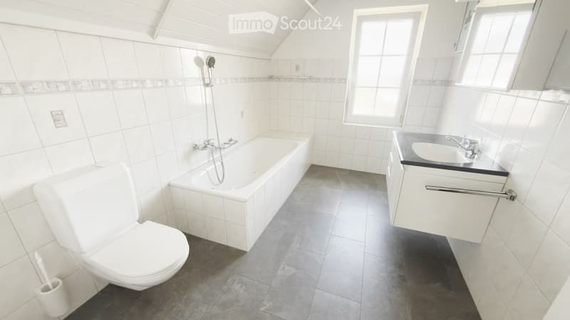 Bright bathroom with white tiles, a bathtub, toilet, sink, and a window letting in natural light.