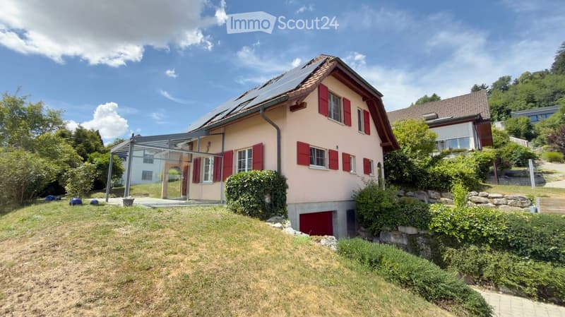 Two-story house with red shutters, solar panels on the roof, and a covered patio area. The house is surrounded by greenery and has a stone wall and driveway.