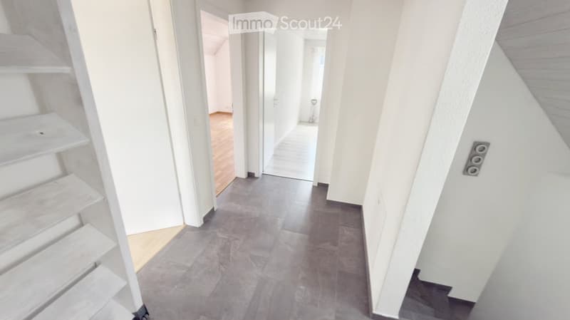 Bright, modern hallway with white walls, grey tiled floor, and a staircase leading upwards. Open doorways lead to other rooms.