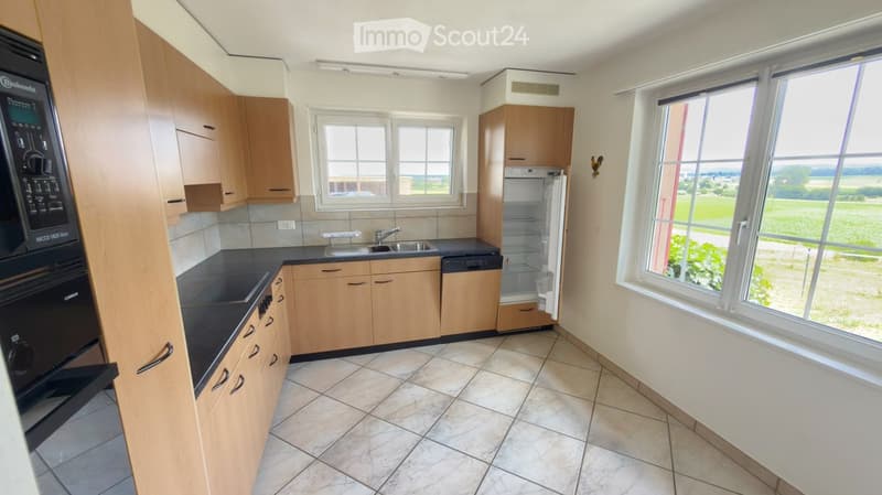 Modern kitchen with light wooden cabinets, black countertops, and stainless steel appliances including a refrigerator, dishwasher, and oven. Large windows offer a view of the countryside.