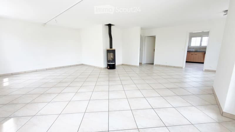 Empty room with tiled floor, white walls, and a central wood-burning stove with a chimney. A doorway leads to another room with cabinets and a sink.