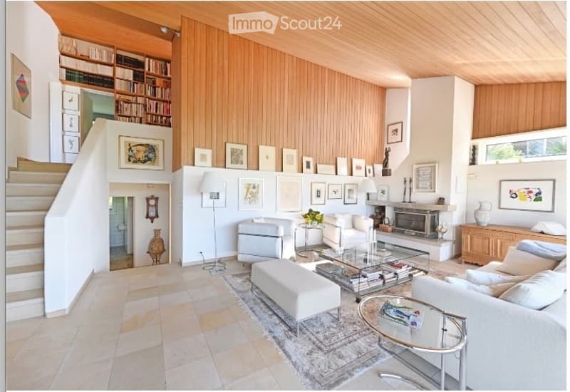 Modern living room with white walls, wooden paneling, and a fireplace. Features a white sofa, ottoman, coffee table, floor lamp, bookshelf, and decorative artwork.