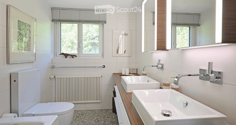 Modern bathroom with white tiles, double sink vanity, toilet, large mirror, and window with blinds. Includes a radiator and decorative picture frame.