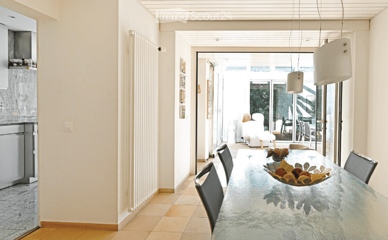 Modern dining area with a glass table, black chairs, and a bowl of fruit. The room has light-colored walls, wooden flooring, and leads to a balcony or terrace area with outdoor furniture.