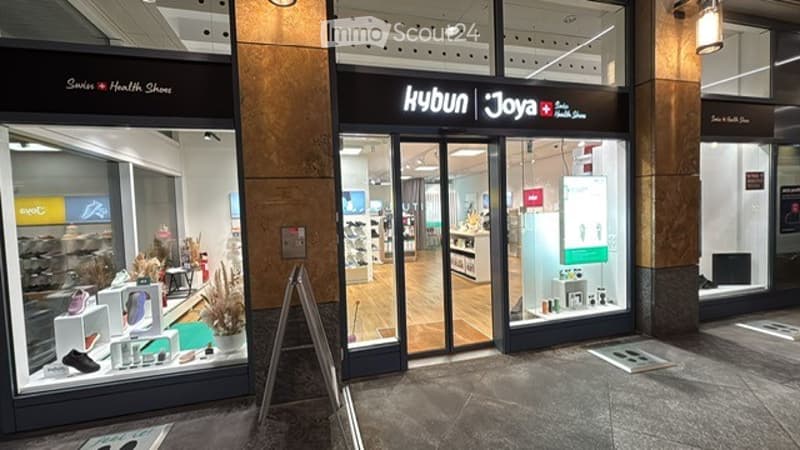 Store with glass entrance, display windows, white shelves, shoes, and various items for sale.
